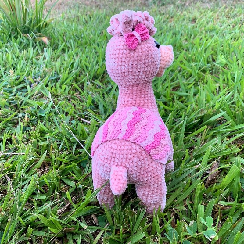Rear view of a small pink crocheted amigurumi animal with a short tail standing on grass