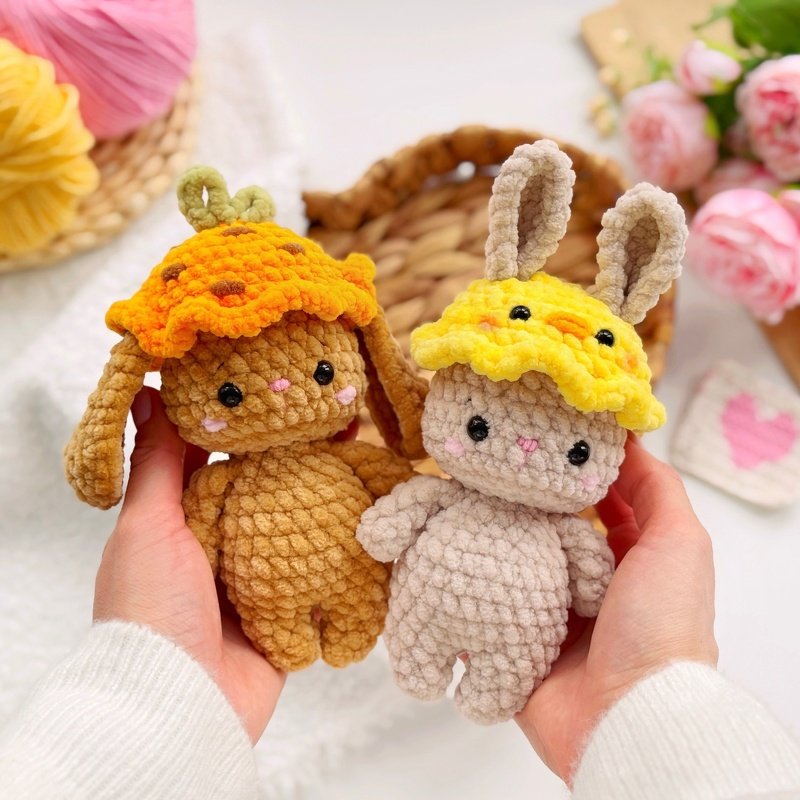 Hands holding two plush crochet amigurumi bunnies wearing yellow duck and orange stem hats