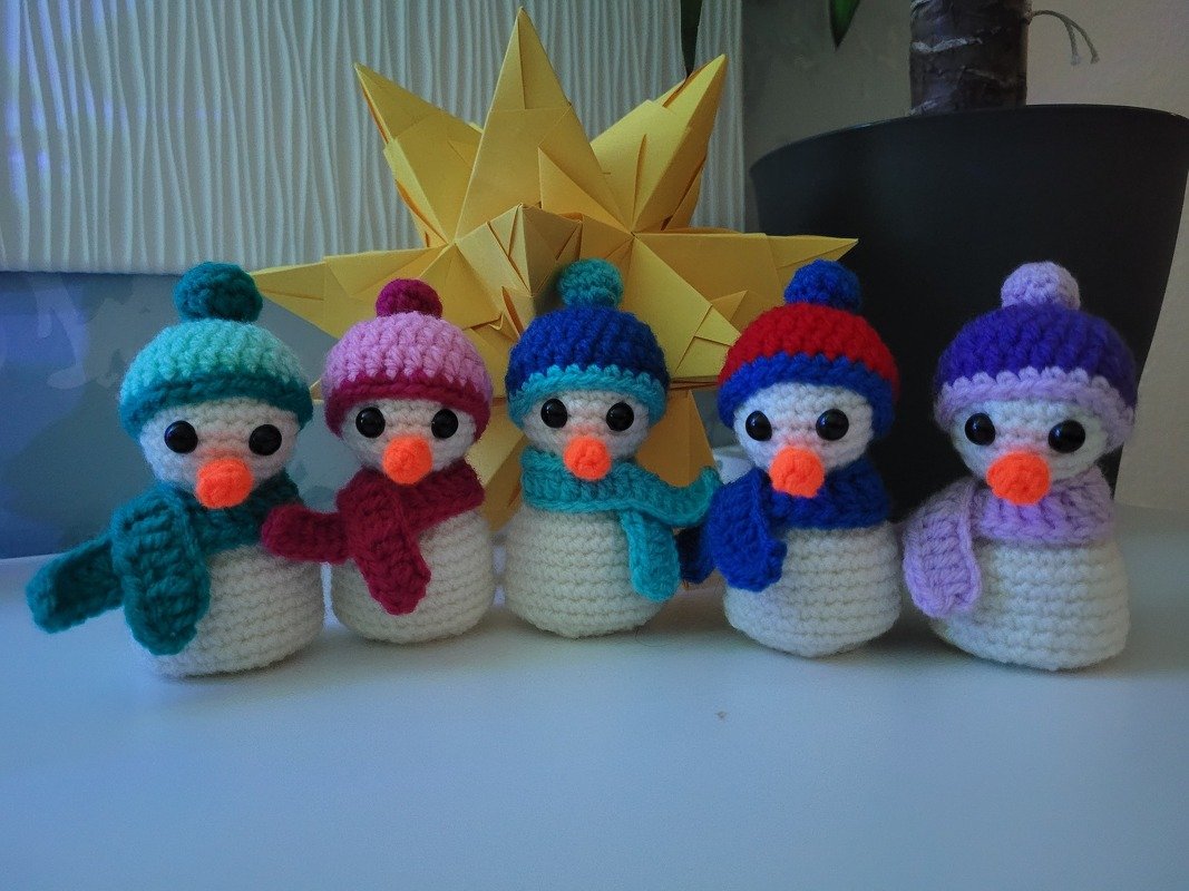 Farbig gehäkelte Schneemänner mit Mützen und Schals vor gelbem Papierstern