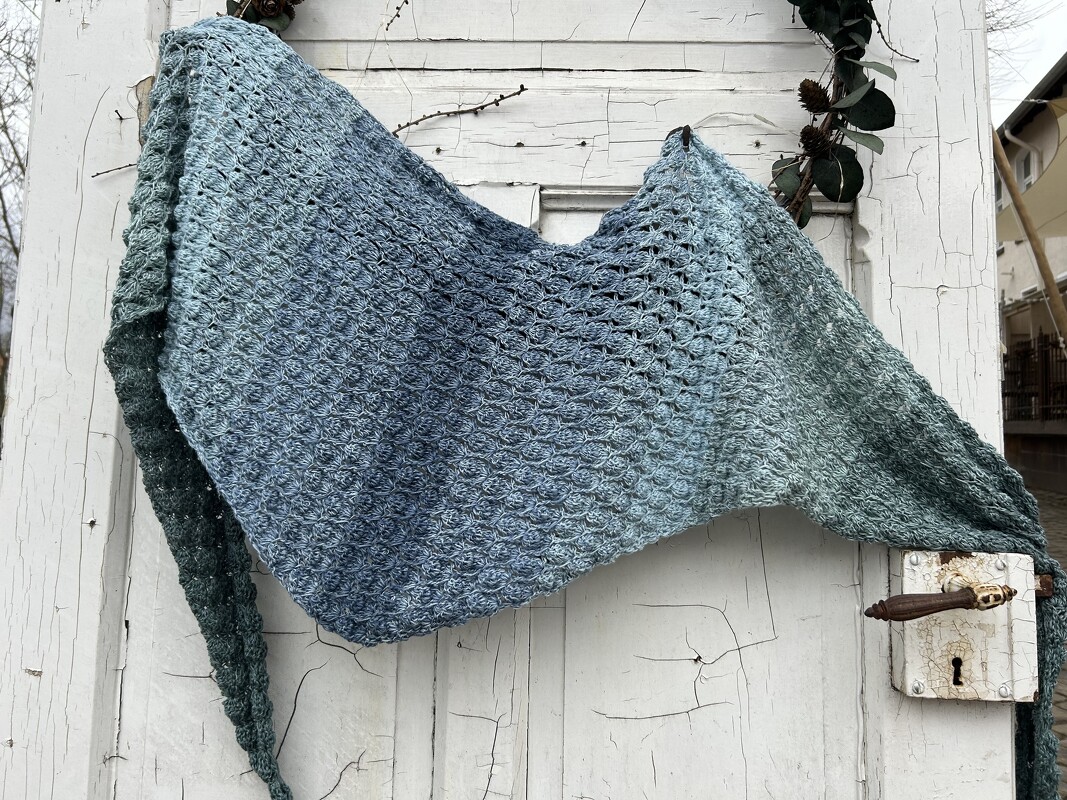 Gehäkeltes Dreieckstuch mit blau-grünem Farbverlauf und Muschelmuster, über einer verwitterten weißen Holztür drapiert