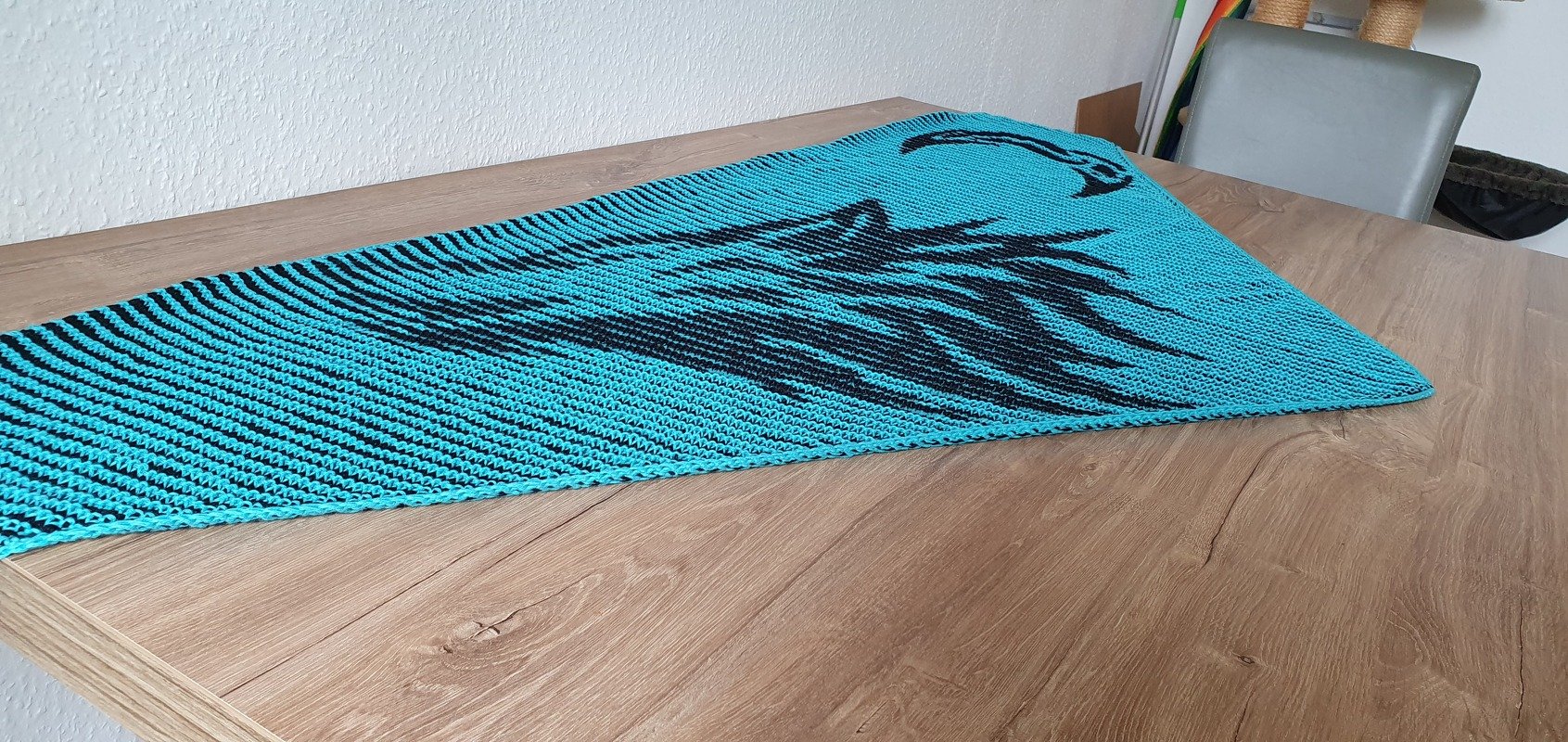 Turquoise garter-stitch triangular shawl with black horse silhouette motif laid flat on a wooden table