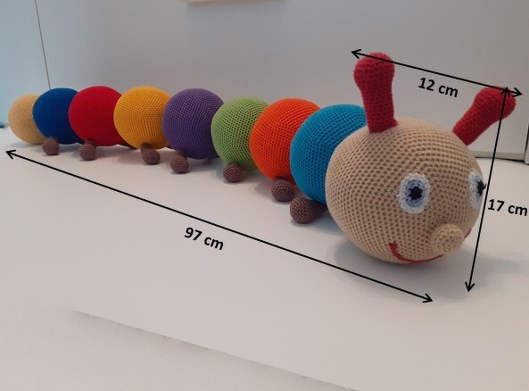 Gehäkelte, bunte Amigurumi‑Raupe mit beige Kopf, blauen Augen, roten Fühlern und mehreren farbigen Körpersegmenten.