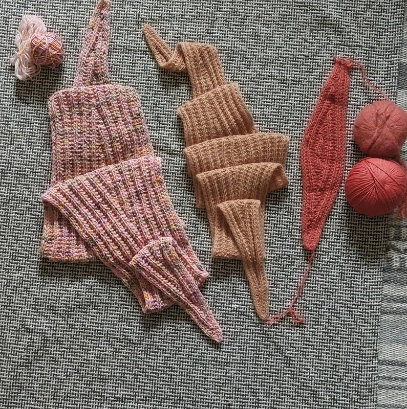 Three ribbed knitted triangle shawl pieces in pink, beige and coral with yarn balls on textured fabric.