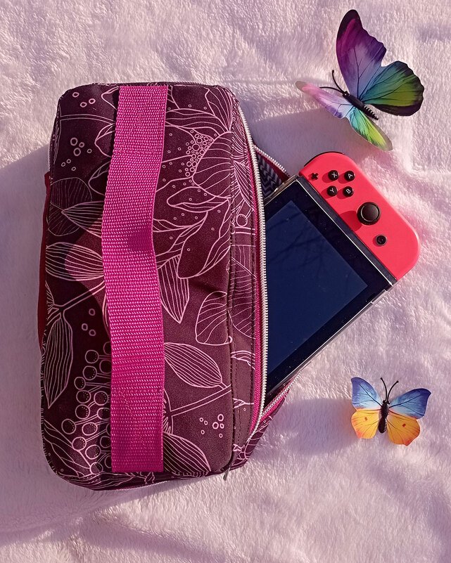 Dunkelviolette Stofftasche mit weißem Floraldruck und pinkem Gurt, daneben rote Handheld-Spielkonsole und bunte Schmetterlingsdeko auf rosa Hintergrund.