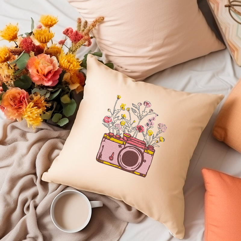 Beiges Kissen mit aufgestickter rosa Kamera und bunten Blumen, daneben ein Blumenstrauß und eine Tasse.