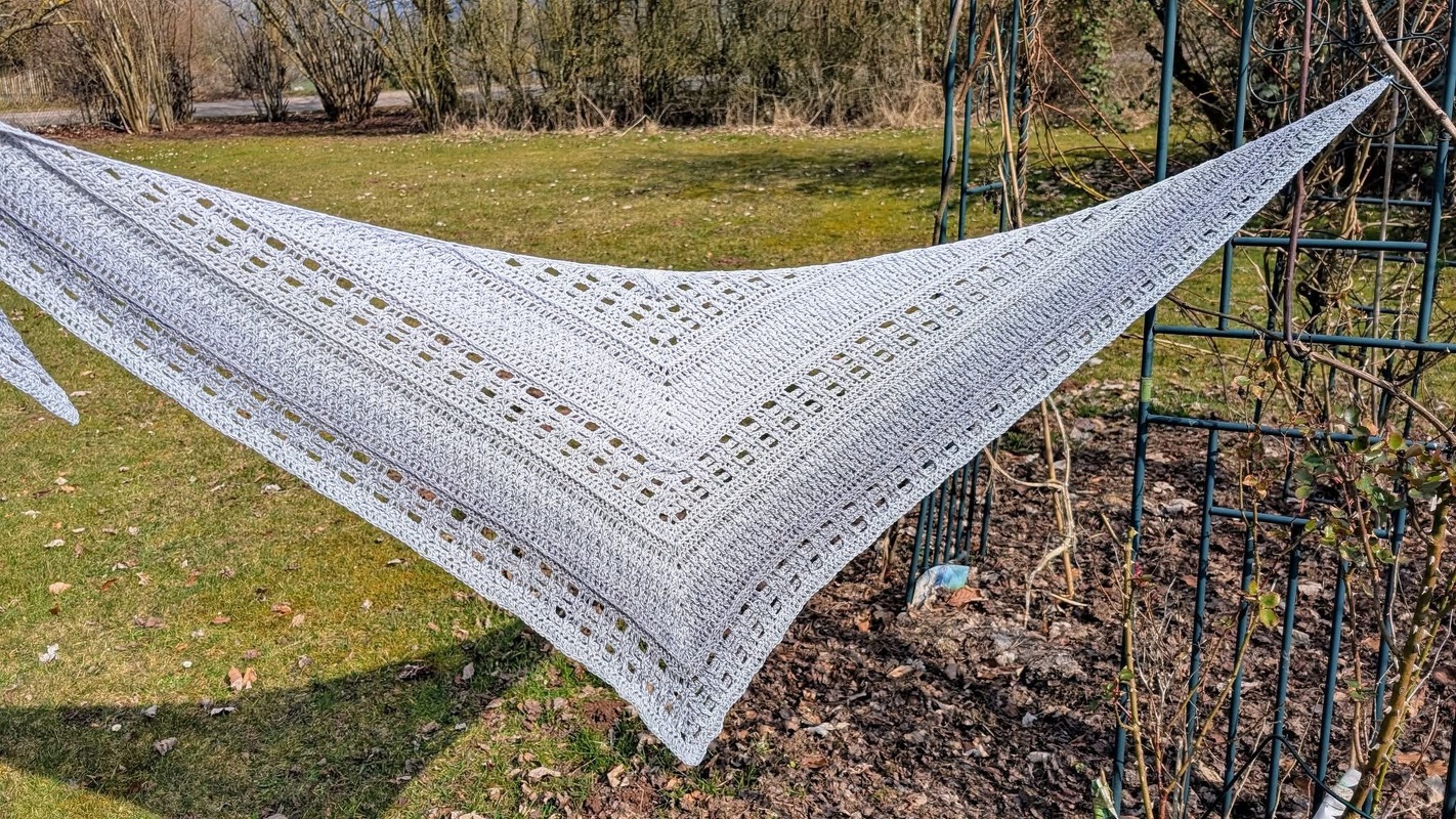 Pale grey crochet triangle shawl with alternating solid bands and lacy square rows stretched on a metal trellis in a garden.