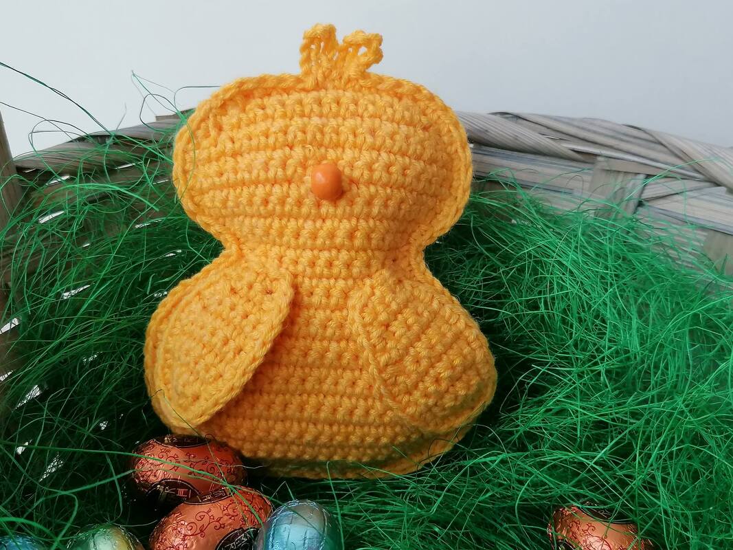 Gehäkeltes gelbes Küken mit orangefarbenem Schnabel in einem Korb mit grünem Gras und bunten Ostereiern.
