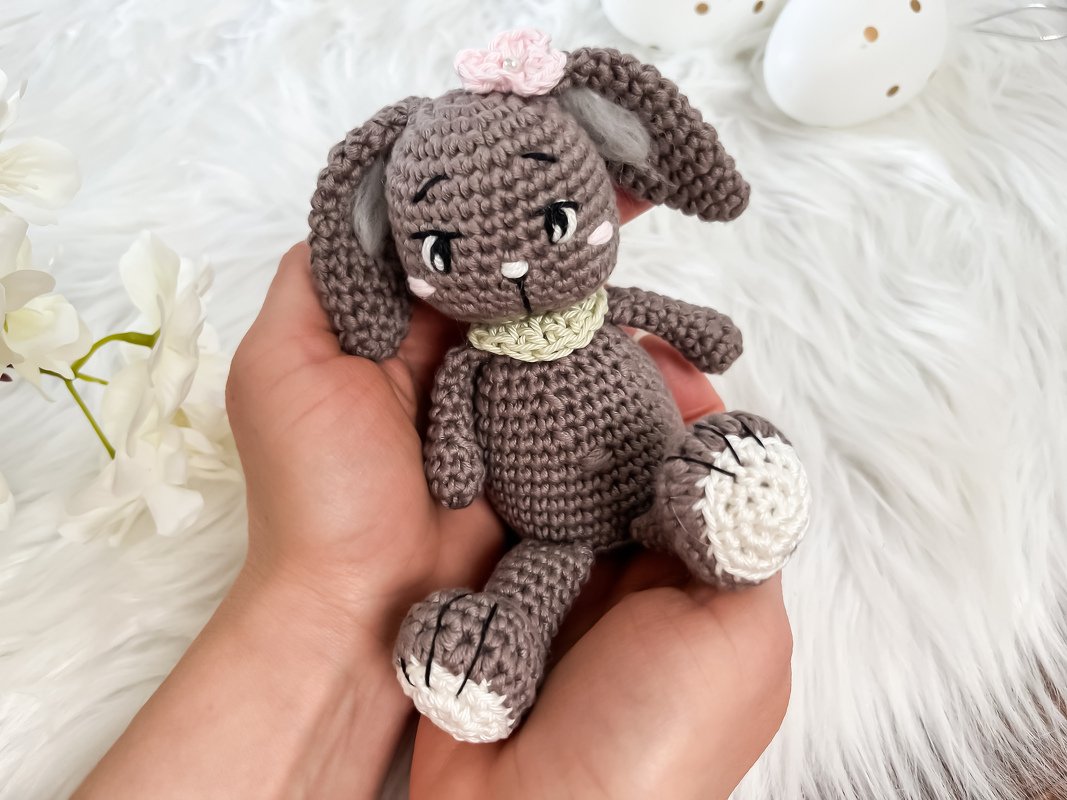 Handgroßes gehäkeltes Amigurumi-Häschen aus grauem Garn mit weißen Pfoten und Blüte am Kopf.