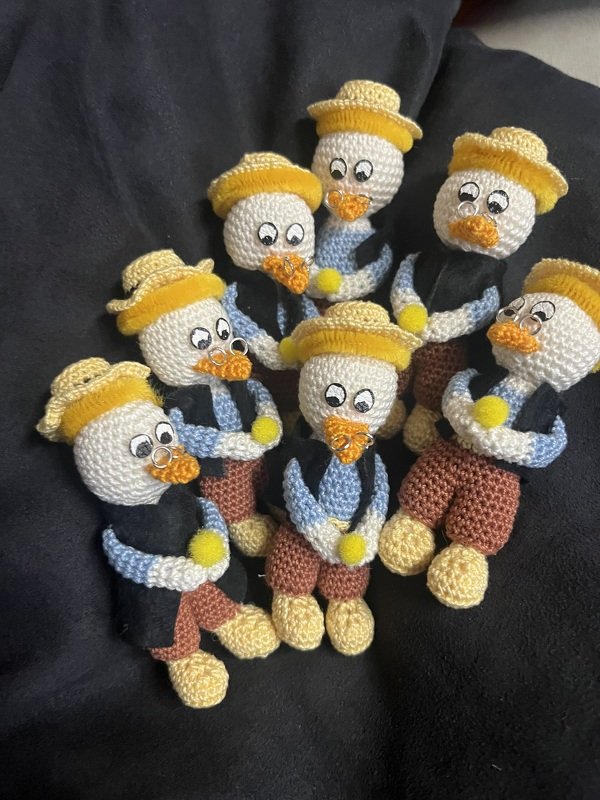 Sieben gehäkelte Entchen-Amigurumi mit gelben Hüten, orangefarbenen Schnäbeln mit Piercings und blauen Armen auf dunklem Hintergrund