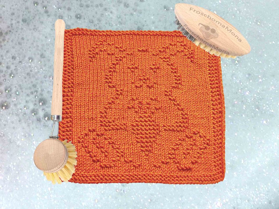 Orangefarbener gestrickter Spüllappen mit Hasenrelief und zwei Holzbürsten auf schaumigem Untergrund.