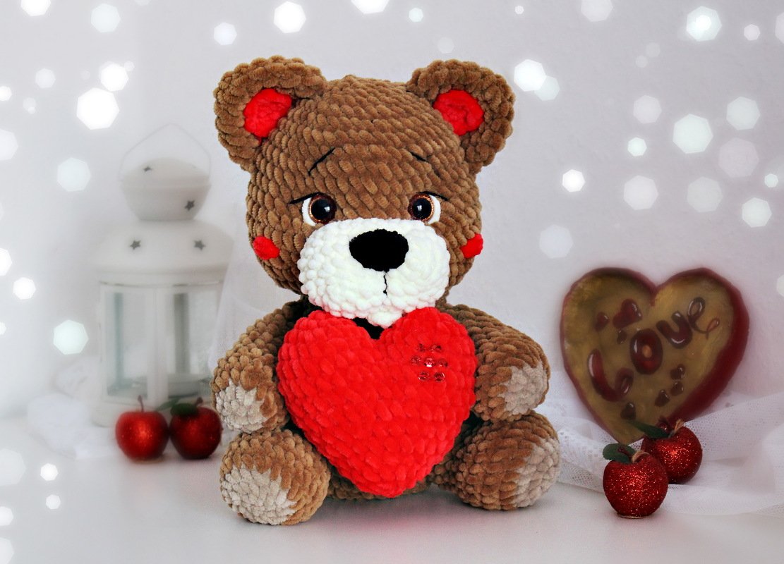 Brauner gehäkelter Amigurumi-Teddybär aus Plüschgarn, sitzend und hält ein rotes Herz vor heller Dekoration mit Lichtern.