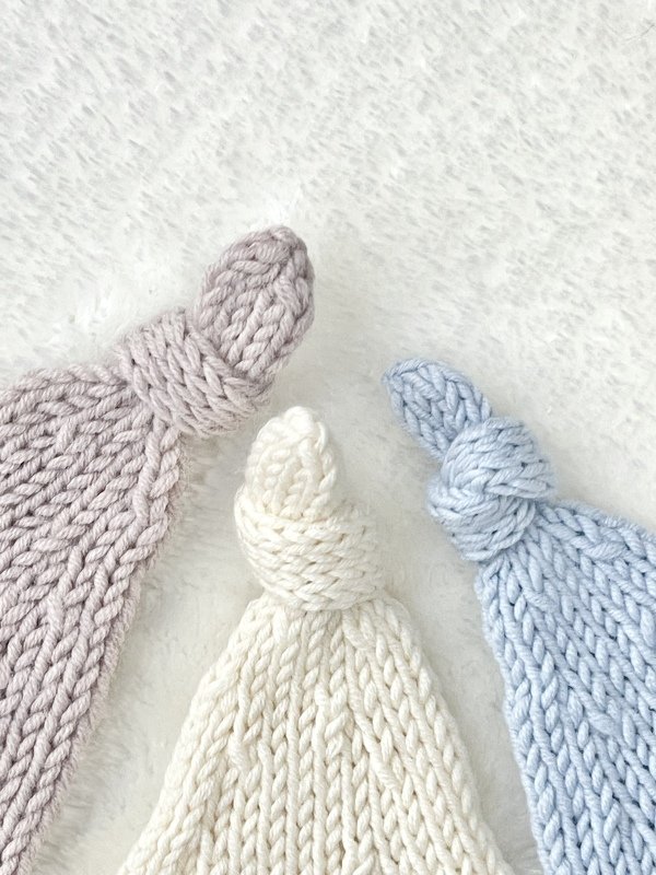 Three knitted baby hats in light grey, cream, and baby blue with top knots on a soft white background.