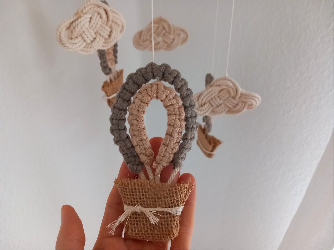 Nahaufnahme eines handgeknüpften Makramee-Heißluftballons mit Jutekorb, von einer Hand gehalten, Teil eines Mobiles mit Wolken im Hintergrund.