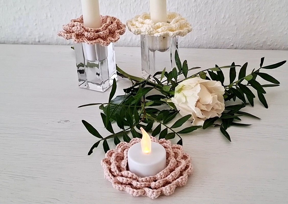 Nahaufnahme von zwei Kerzenständern mit gehäkelten Kerzenringen in Rosa und Creme, einem rosa gehäkelten Untersetzer mit LED-Teelicht und einer weißen Rose mit Grün auf einem weißen Holztisch.
