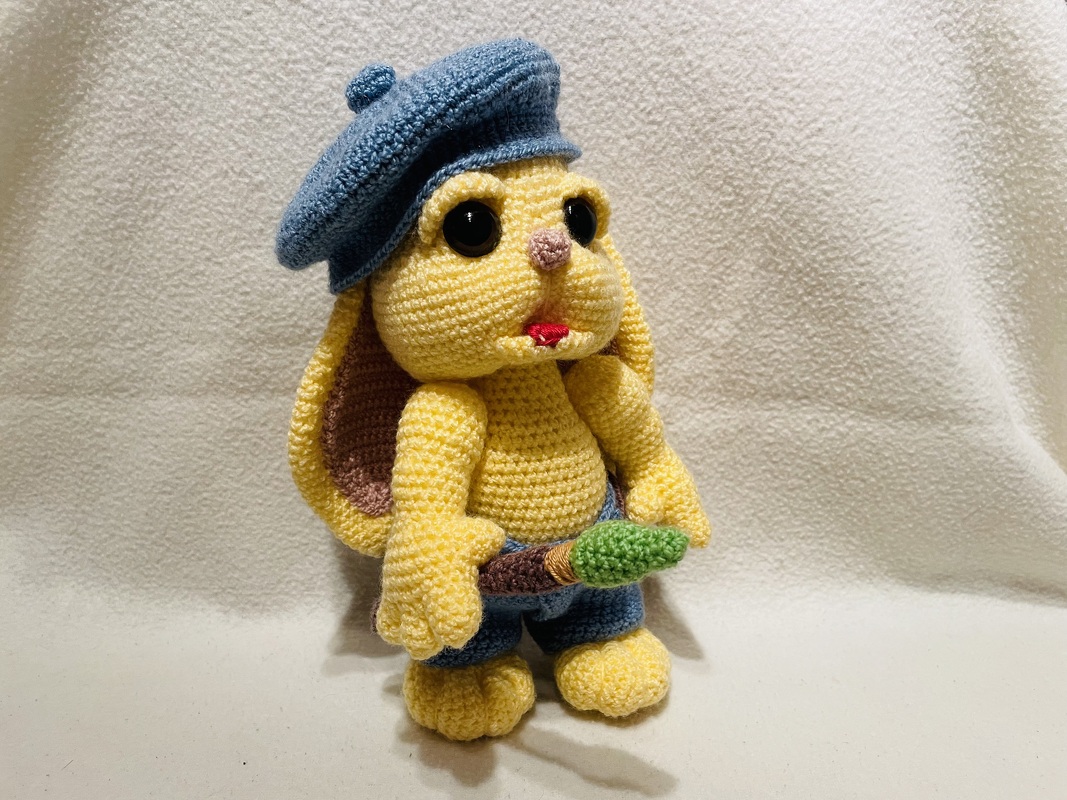 Gelber gehäkelter Hase mit blauer Baskenmütze, blauer Hose und Karotte