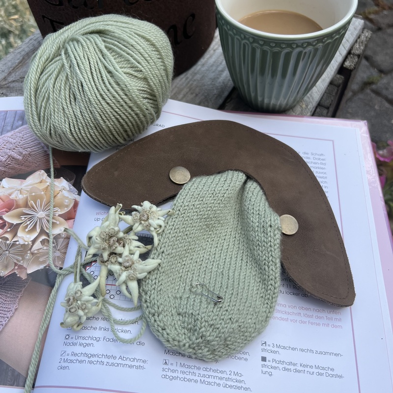 Gestricktes Taschenteil aus hellgrüner Wolle mit Lederlasche, Edelweißblüten und Kaffeetasse auf Strickmusterbuch