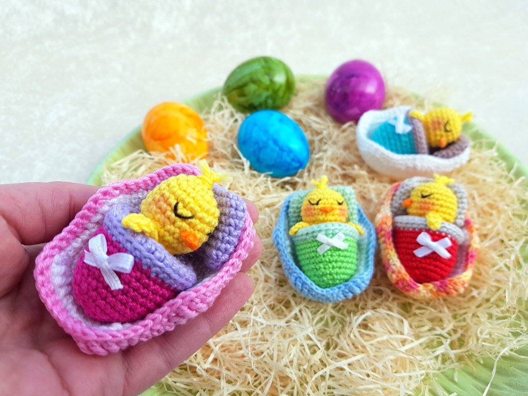 Hand hält ein pinkes gehäkeltes Nest mit schlafendem gelbem Mini-Küken; im Hintergrund weitere bunte gehäkelte Nestchen und gefärbte Eier auf Stroh.