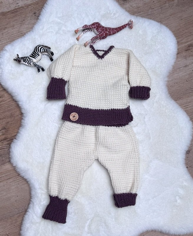 Gehäkeltes Kinder-Set aus Pullover mit V‑Ausschnitt und passender Hose in Creme mit dunkellila Bündchen, auf weißem Fell mit Spielzeuggiraffe und Zebra drapiert