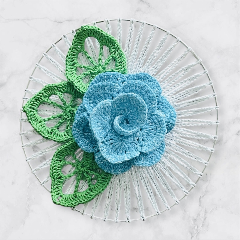 Gehäkelte blaue Blume mit drei grünen Blättern auf einem weißen, mit Garn umwickelten Dekoring auf marmoriertem Hintergrund.