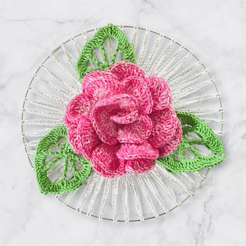 Gehäkelte pinke Rose mit drei grünen Blättern auf einem runden Metallring mit weißem Wickelgarn