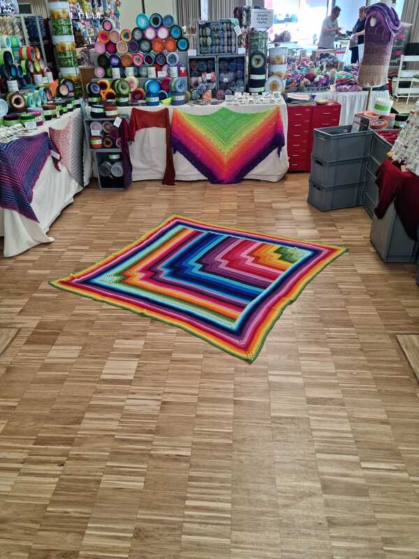 Quadratische Häkeldecke in leuchtenden Regenbogenfarben auf Holzfußboden, im Hintergrund Garnstände
