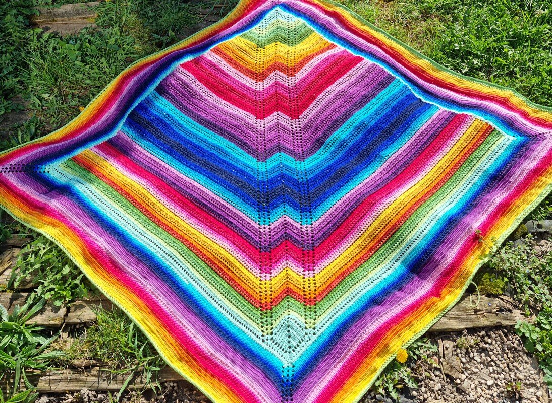 Gehäkelte Decke mit Zickzackmuster in Regenbogenfarben auf Gras und Holzplanken