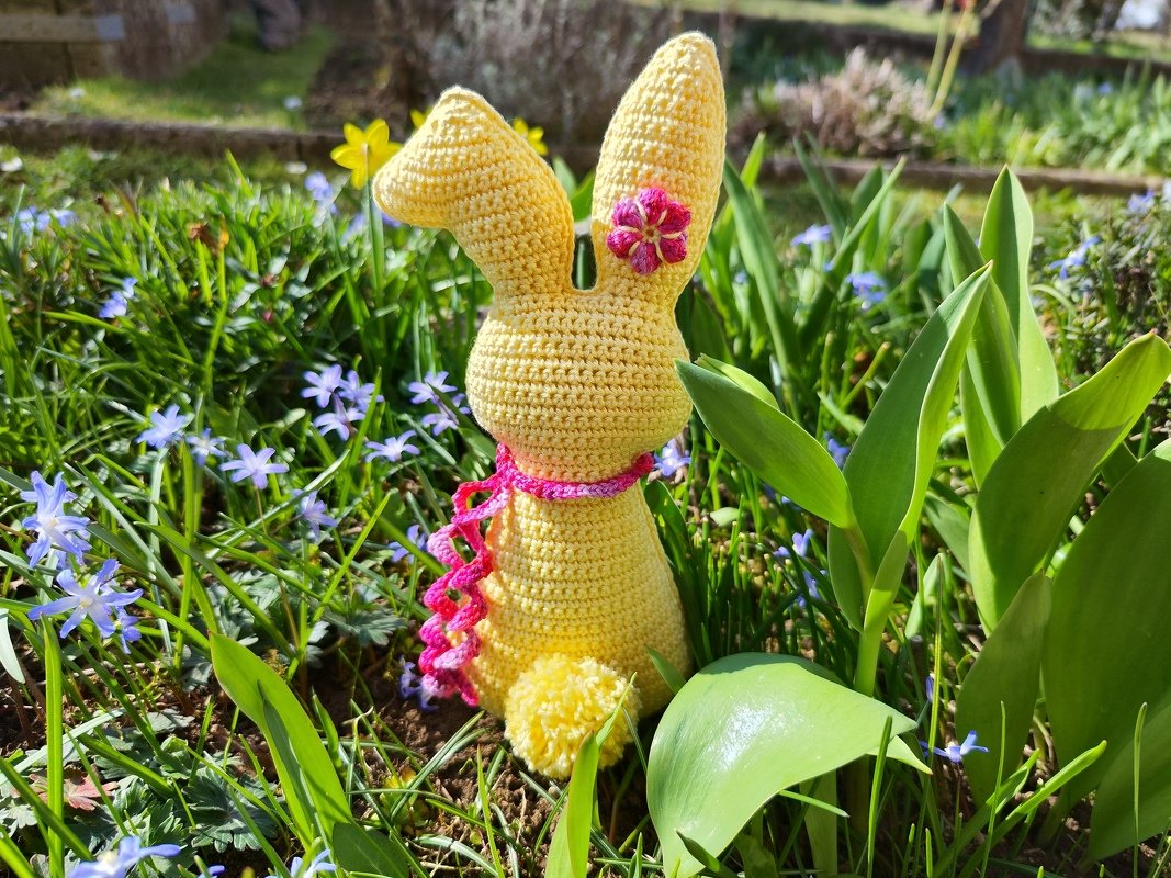 Gelber gehäkelter Amigurumi-Hase mit Blume am Ohr und pinkem Spiralschal im Garten