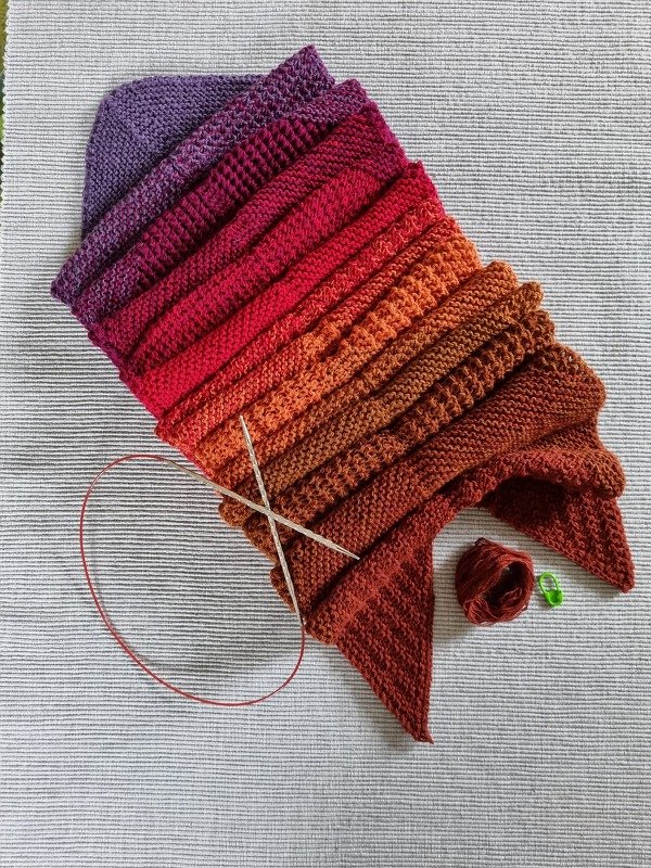 Halbfertiger gestrickter Schal mit Farbverlauf von Lila über Rot bis Orange-Braun, Texturstreifen, Rundstricknadeln und Garnknäuel auf hellem Untergrund