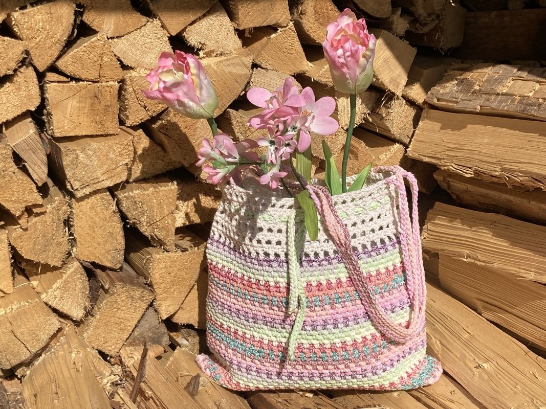 Gehäkelte Umhängetasche in Pastellfarben mit Blumen vor gestapeltem Kaminholz