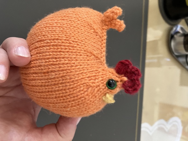 Hand hält ein orangefarbenes, handgestricktes Amigurumi-Huhn mit rotem Kamm und grünen Augen.