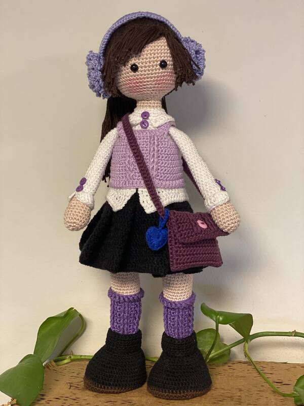 Hand-crocheted amigurumi doll in lavender vest, earmuffs, black skirt and boots, carrying a small purple crossbody bag.