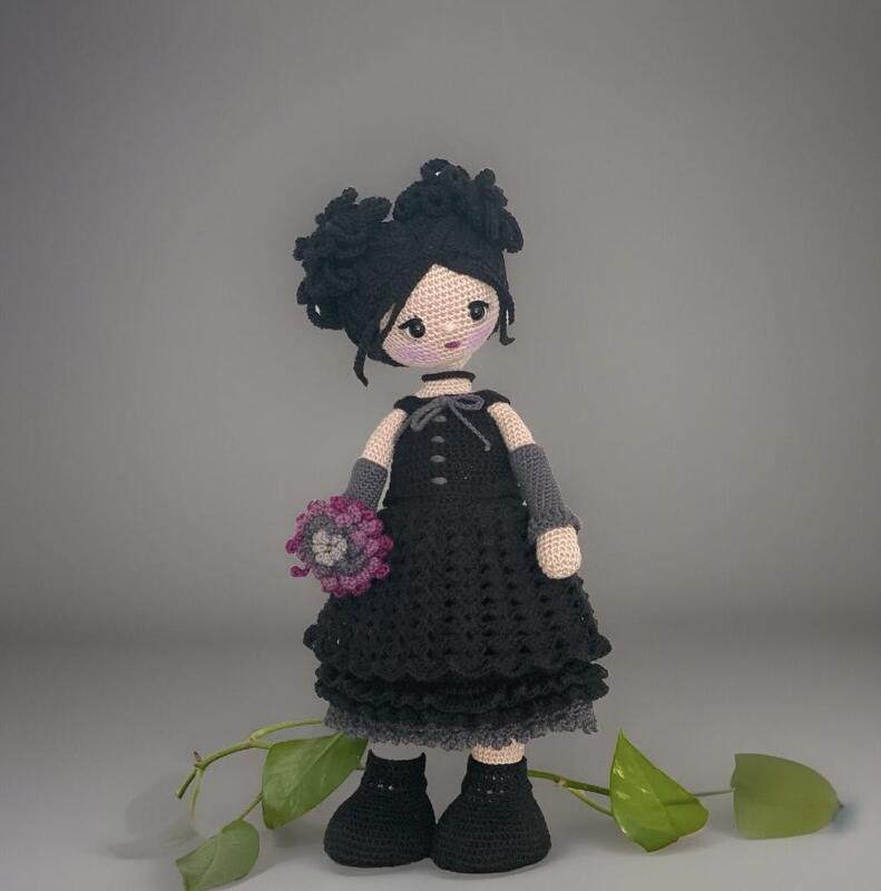 Crocheted amigurumi doll wearing a black dress, grey fingerless gloves, and black crocheted shoes, holding a small purple and grey flower bouquet.