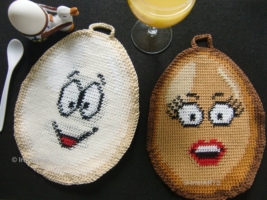 Nahaufnahme von zwei gehäkelten eiförmigen Topflappen mit unterschiedlichen Gesichtsmotiven, liegend neben einem Eierbecher, einem weißen Löffel und einem Glas Saft auf dunklem Untergrund.