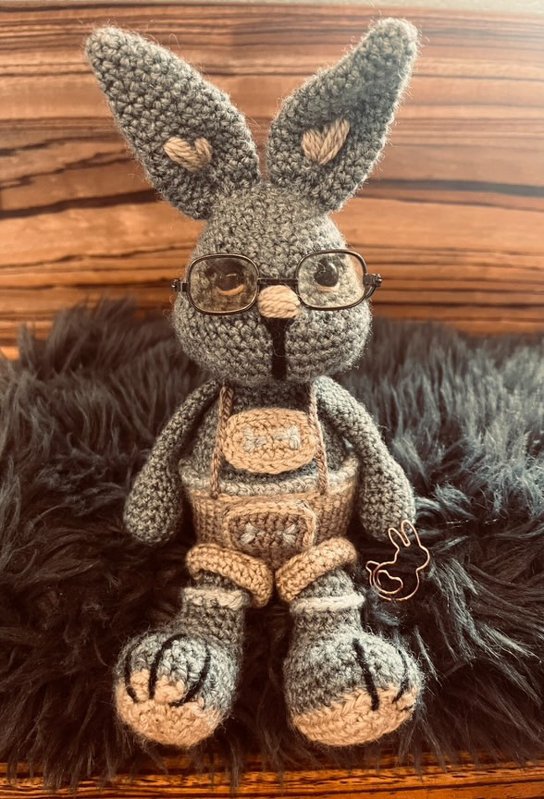 Gehäkelter grauer Amigurumi‑Hase mit Brille und beigen Lederhosen auf dunklem Fell