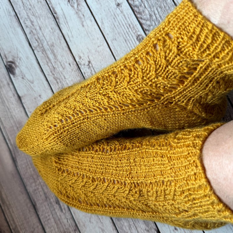 Ein Paar gestrickte Damensocken in Senfgelb mit detailliertem Ajourmuster auf Holz.