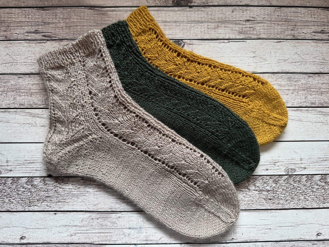 Drei gestrickte knöchelhohe Damensocken in Beige, Dunkelgrün und Senfgelb mit feinem Ajourmuster, nebeneinander auf hellem Holzhintergrund.