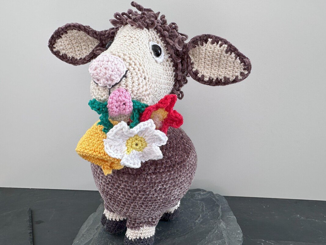 Gehäkeltes Amigurumi-Schaf mit braunem Körper und hellbeigem Kopf, geschmückt mit Blumenkranz und gelber Glocke.