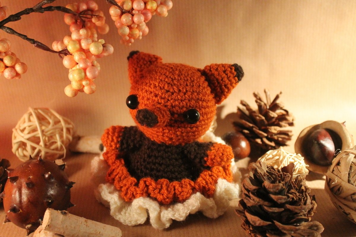 Gehäkelter orange-brauner Amigurumi-Fuchs mit schwarzen Sicherheitsaugen, umgeben von Tannenzapfen, Kastanien und Deko-Kugeln.