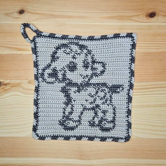 Quadratischer gehäkelter Topflappen mit Lämmchen-Motiv in Hell- und Dunkelgrau und Aufhängeschlaufe auf Holzuntergrund.