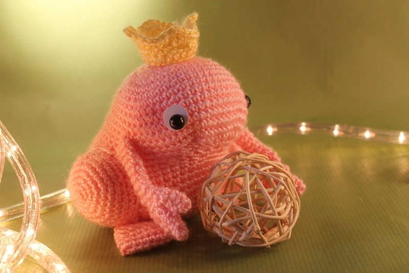 Rosafarbener gehäkelter Oktopus mit gelber Krone, Wackelaugen und runder Bastkugel vor grünem Hintergrund