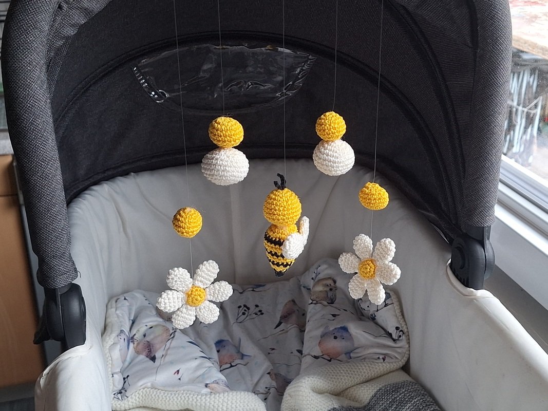 Handmade crocheted bee, daisies and yellow-and-white balls mobile hanging inside a grey stroller bassinet.