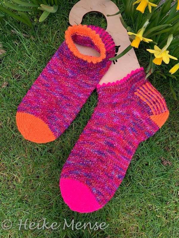 Kurzschaftige, handgestrickte Socken in Pink, Magenta und Orange auf Gras neben Narzissen und einem Sockenbrett