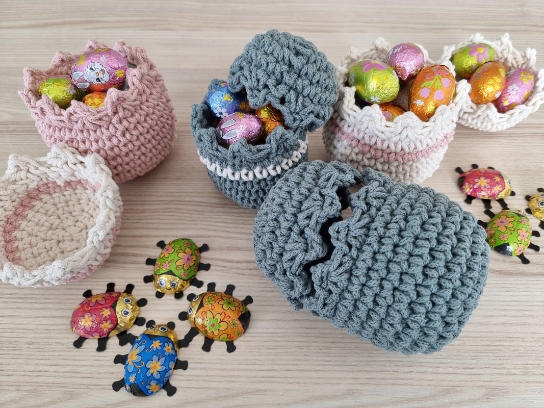Mehrere gehäkelte kleine Körbchen in Rosa, Hellbeige und Grau, gefüllt mit bunten Schokoladeneiern und Schokoladen-Marienkäfern auf hellem Holztisch.