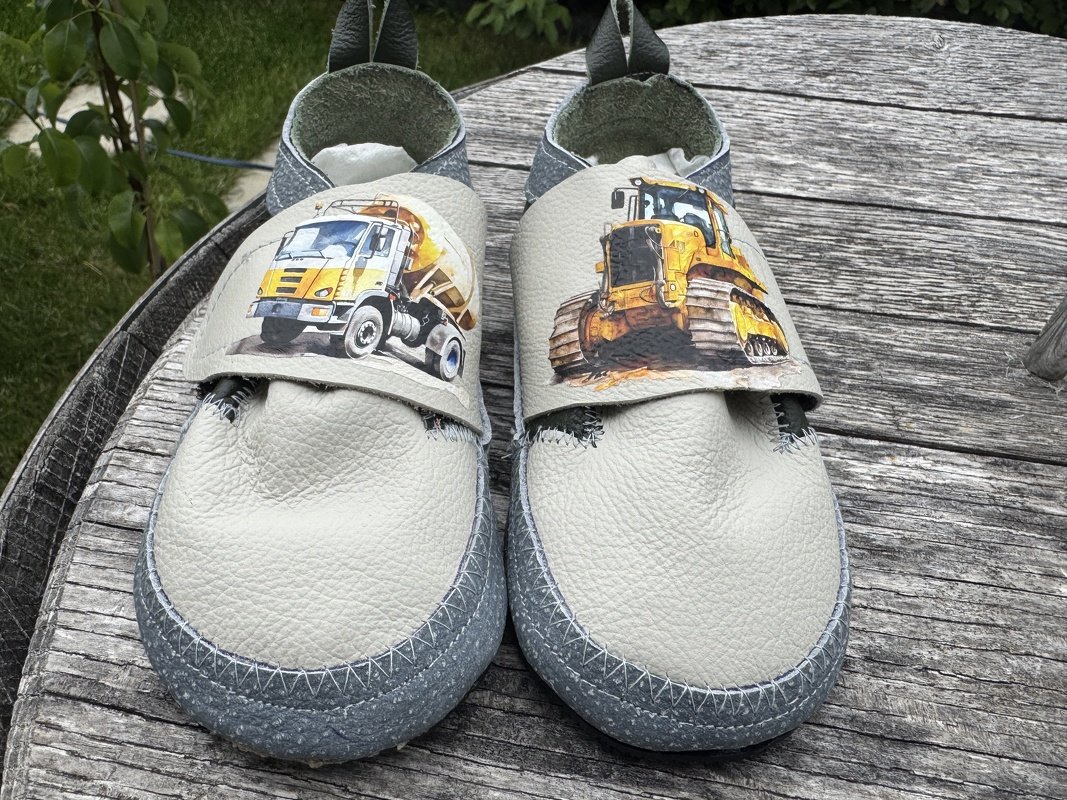 Hellgraue Kinderpuschen aus Leder mit Prints eines Betonmischers und eines Raupenladers auf einer Holzplatte.
