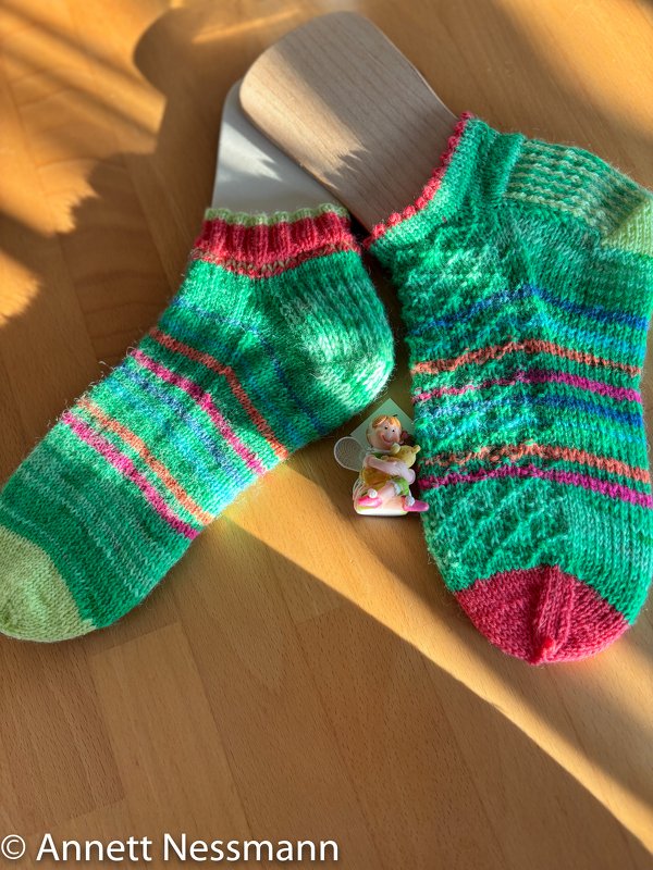 Zwei kurzgestrickte grüne Socken mit rosa und blauen Streifen auf Holz-Sockenspannern, liegend auf einem Holztisch, daneben eine kleine Figur