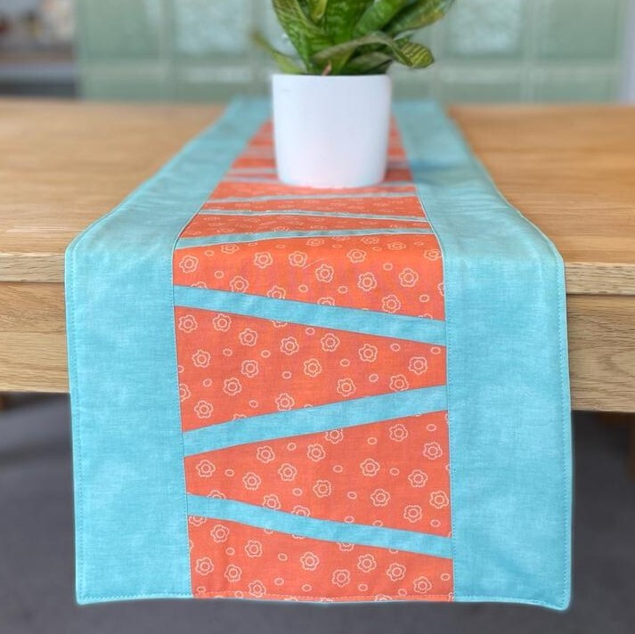 Tischläufer mit türkisfarbenem Rand und orangefarbenem Mittelteil mit diagonalen Patchworkstreifen, auf Holztisch mit Pflanzentopf