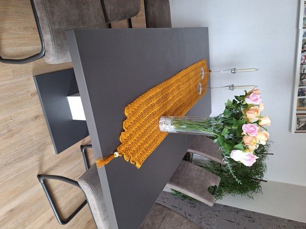 Senfgelber gehäkelter Tischläufer mit Muschelrand und Quasten auf dunkelgrauem Esstisch, Glasvase mit rosa Rosen und Kerzenhaltern