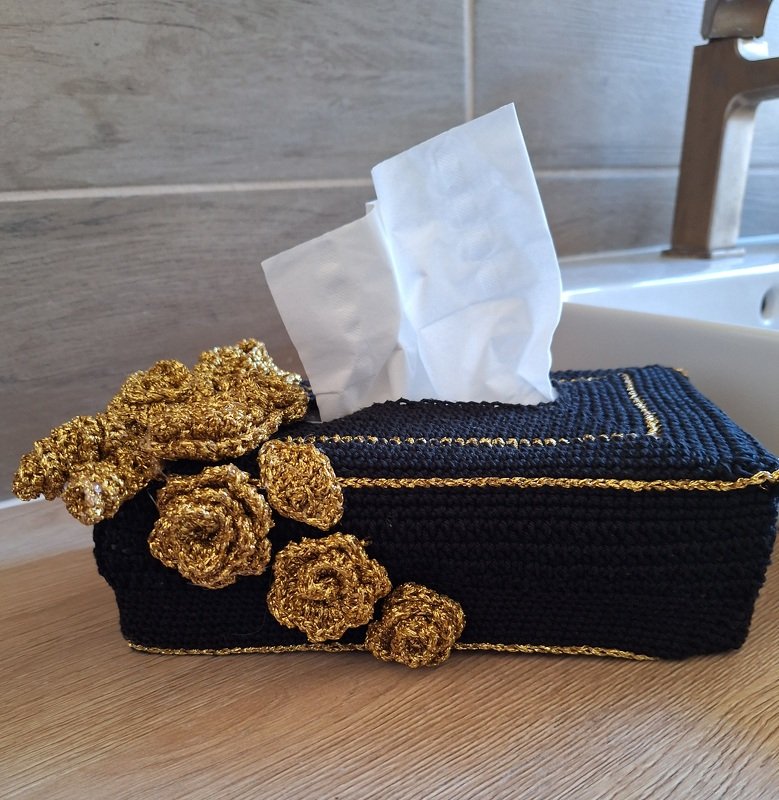 Gehäkelte schwarze Taschentuchbox mit goldenen Rosenapplikationen und goldener Umrandung, auf einer Holzoberfläche neben einem Waschbecken.