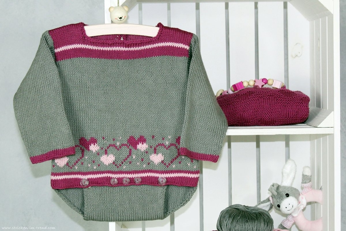 Gestrickter Babystrampler in Graugrün mit rosa Herzbordüre auf einem Regal