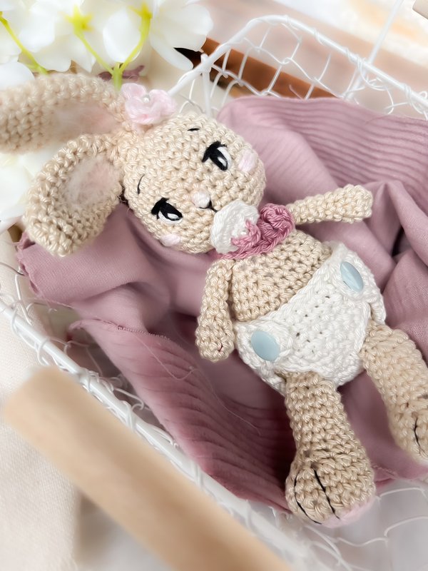 Gehäkeltes Amigurumi-Häschenbaby mit Schnuller und Windelhose im Drahtkörbchen auf rosa Decke